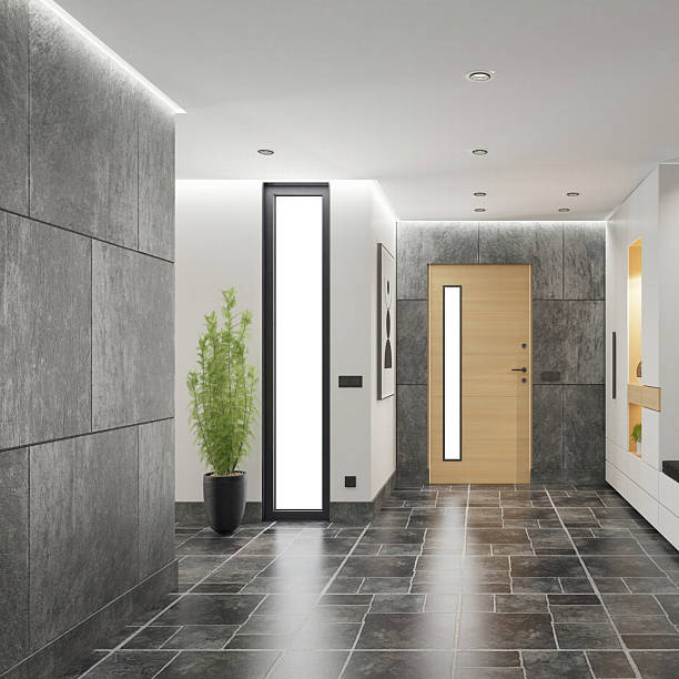 Modern building entrance hallway design featuring dark stone tile flooring and a mix of smooth white and textured gray walls. Light wood door with vertical frosted glass panel is centered, flanked by a tall window and potted plant. Built-in white cabinet with warm lighting and mirror adds depth. Abstract wall art and recessed ceiling lights complete the clean, modern look. Ideal for interior design, architecture, lifestyle, and residential themes.