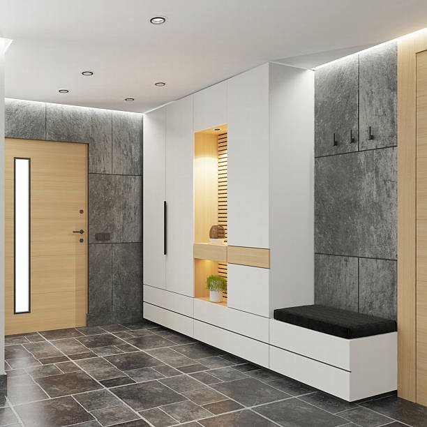 Modern building entrance hallway design featuring dark stone tile flooring and a mix of smooth white and textured gray walls. Light wood door with vertical frosted glass panel is centered, flanked by a tall window and potted plant. Built-in white cabinet with warm lighting and mirror adds depth. Abstract wall art and recessed ceiling lights complete the clean, modern look. Ideal for interior design, architecture, lifestyle, and residential themes.